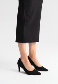 Czarne zamszowe szpilki z ostrym noskiem i zwężającym się obcasem. Charakteryzują się gładką teksturą i prostym designem. Nosi się je z prostą czarną spódnicą.