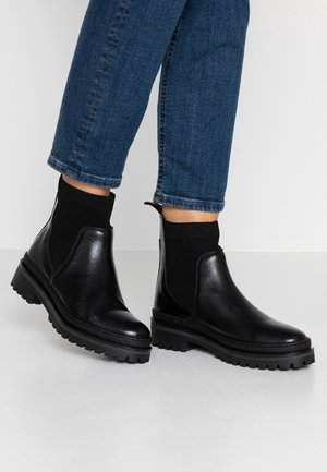 Musta nahkasaappaat nilkkapitkällä, tekstuurilla varustetulla pinnalla, elastisella sukka-kaltaisella yksityiskohdalla ja karkealla kumipohjalla. Yhdistetty sinisiin farkkuihin.