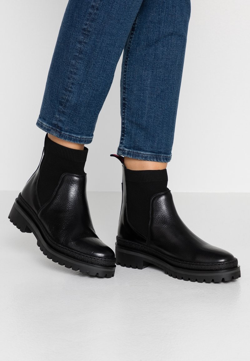 Musta nahkasaappaat nilkkapitkällä, tekstuurilla varustetulla pinnalla, elastisella sukka-kaltaisella yksityiskohdalla ja karkealla kumipohjalla. Yhdistetty sinisiin farkkuihin.