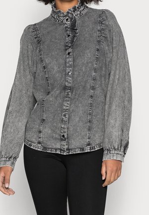 Woman wearing a washed black denim blouse with ruffled high collar and snap buttons, paired with black pants.