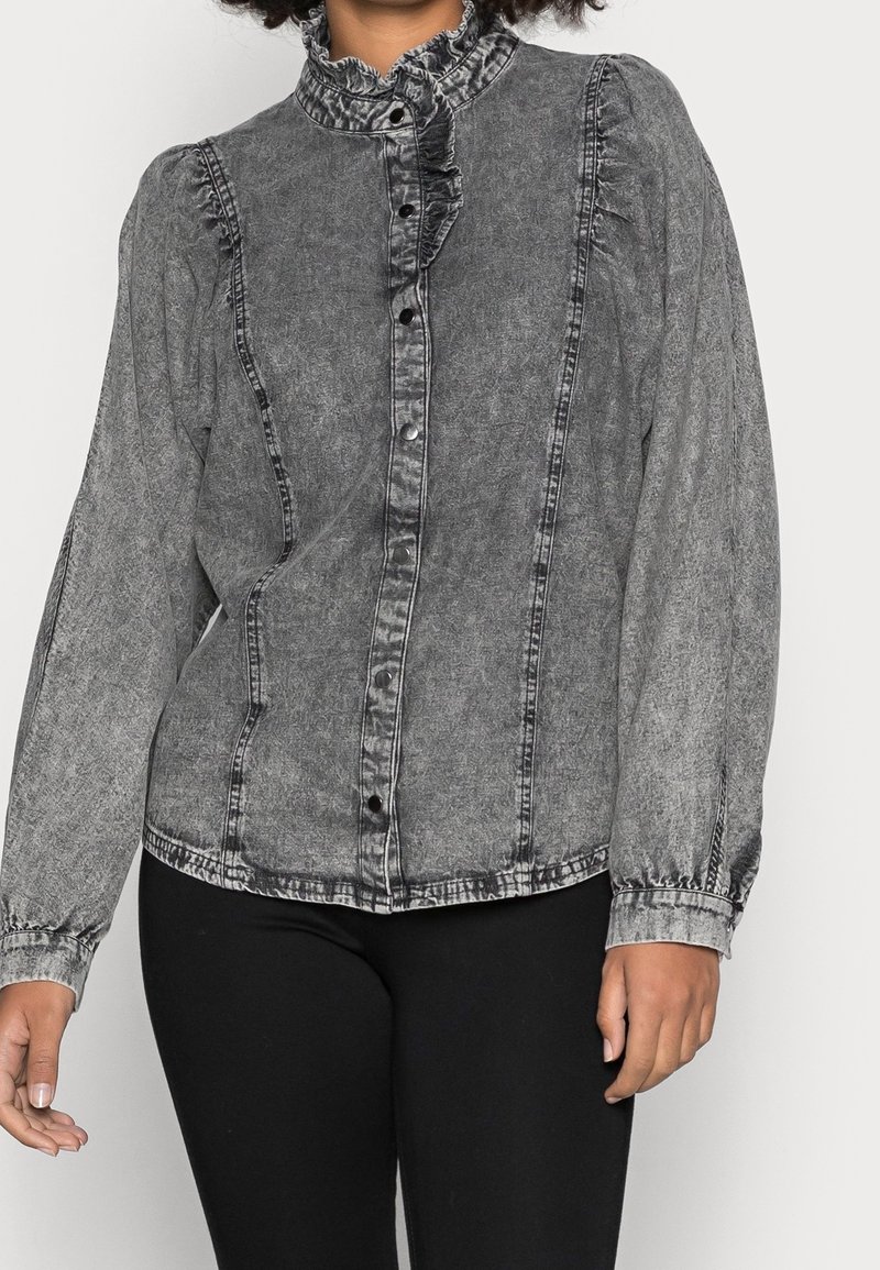 Kvinna i en tvättad svart denimskjorta med volang-krage och tryckknappar, parat med svarta byxor.