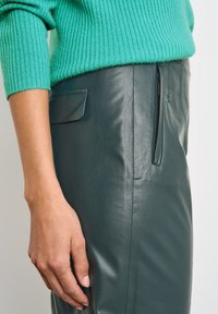 Ruka odpočívá vedle tmavě zelených kožených kalhot se zipem a klopovou kapsou, nošených s hnědým svetr s ribováním ve vodní modré barvě.