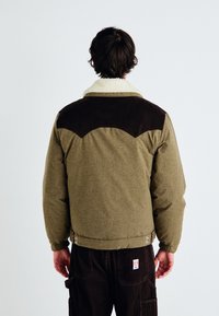 Veste vert olive avec un empiècement en velours côtelé marron, un col en fourrure synthétique et des poignets élastiques. Présente une texture lisse et des pressions en bas.