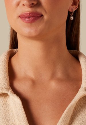 Nahaufnahme des unteren Gesichtsbereichs und Hals einer Frau, die einen beigen, strukturierten Kragenpullover und einen silbernen Creolen-Ohrring mit einem hängenden Edelstein trägt.