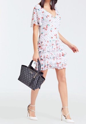 Femme portant une robe à fleurs blanche à volants, tenant un sac à main de créateur noir à motif, chaussée de chaussures blanches à talons hauts et bouts pointus.