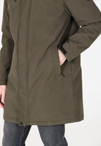 Giacca verde oliva con taglio dritto, dotata di tasche laterali, chiusure in velcro sui polsini e una finitura di tessuto liscia e resistente all'acqua.
