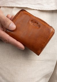 Braune Lederbörse, die in einer Hand gehalten wird; verfügt über einen Reißverschlussverschluss, eine glatte Textur und den eingeprägten Markennamen auf der Vorderseite.