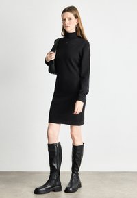 Vestido de malha preto com colarinho alto, mangas longas e um ajuste descontraído. Combinado com botas de couro preto até ao joelho e uma mala de ombro preta.