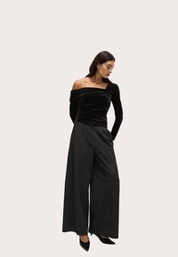 Schwarzes Samt-Oberteil ohne Träger, kombiniert mit hoch taillierten, fließenden schwarzen Hosen. Spitz zulaufende schwarze Schuhe runden das Outfit ab.