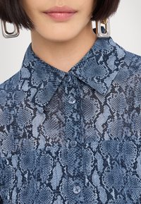 Gros plan d'une personne portant une chemise boutonnée à imprimé serpent bleu et noir avec un col pointu et de grandes boucles d'oreilles géométriques argentées.