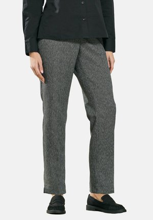 Bas du corps d'une personne portant une chemise à manches longues noire, un pantalon gris texturé à jambes droites et des mocassins noirs sans lacets, debout devant un fond uni.