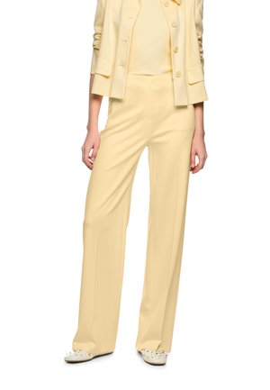 Frau trägt hellgelbe, weit geschnittene Hose, passende Jacke und weiße Schuhe mit Nieten, steht mit entspannt an den Seiten hängenden Händen.