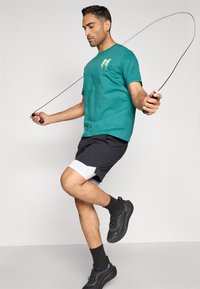 Μπλούζα τιρκουάζ με κοντά μανίκια και κίτρινο μοτίβο, συνδυασμένη με μαύρο αθλητικό σορτς με λευκές λεπτομέρειες. Το μοντέλο πηδά σχοινάκι φορώντας μαύρα αθλητικά παπούτσια.