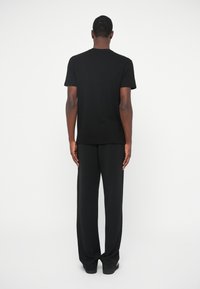 Mees seisab seljaga, seljas tavaline must lühikeste varrukatega t-särk, mustad laiad püksid ja mustad kingad, valge tausta ees.