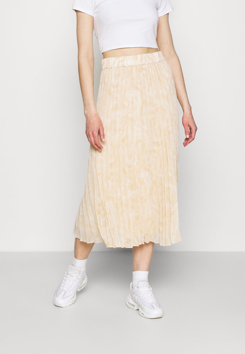 Plisserad midikjol i mjukt, ljust beige tyg med texturerad yta och resår i midjan, kombinerad med vita sneakers och strumpor.