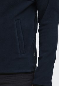 Marineblå fleecejakke med sidelomme, strukturert stoff og en tettsittende design med svarte detaljer ved kantene og ermer.