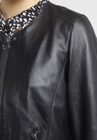 Černá kožená bombardérka s hladkým povrchem, vybavená vysokým límcem se vzorem černobílých skvrn a zipovým zapínáním.