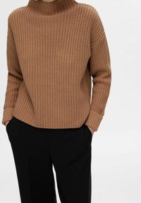 Pull marron en maille avec un col montant et des poignets côtelés, assorti à un pantalon noir. La texture est épaisse et la coupe est décontractée.