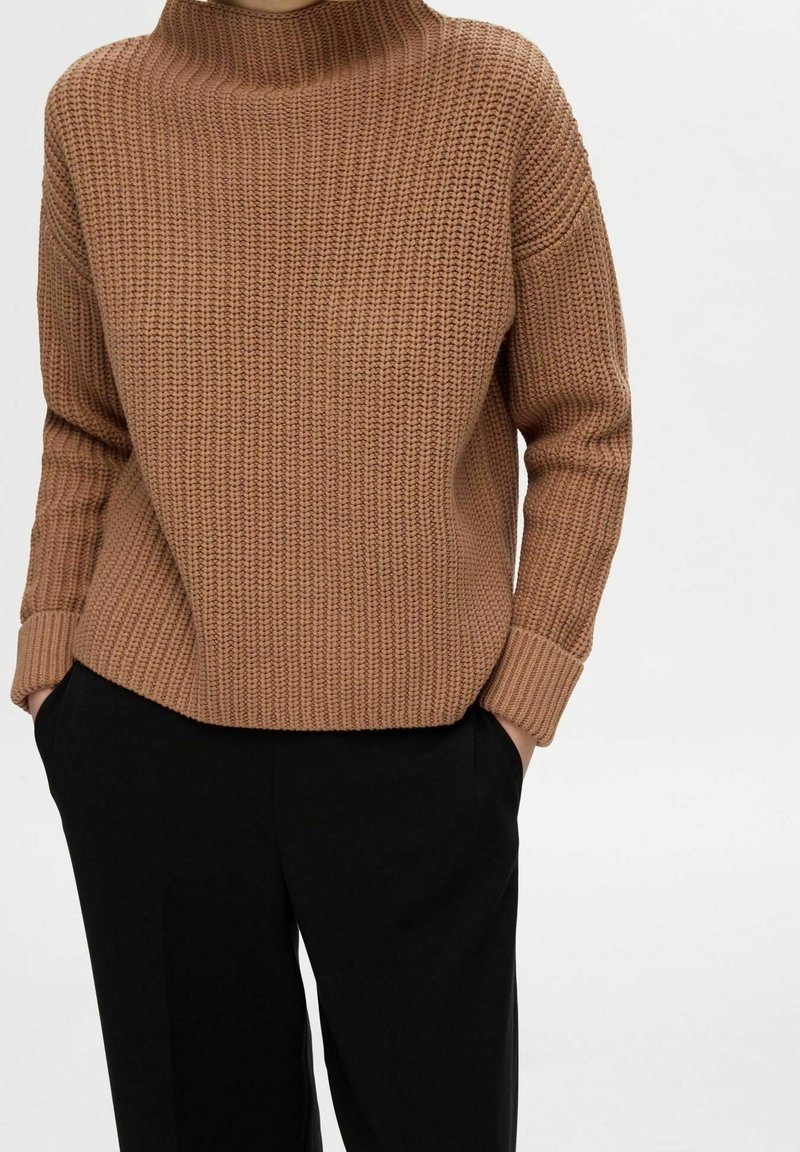 Pull marron en maille avec un col montant et des poignets côtelés, assorti à un pantalon noir. La texture est épaisse et la coupe est décontractée.