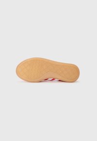 Gummisula i beige med en zigzag-mönstrad yttersula, med röda och vita ränder. Platt, rund form med en slät yta.