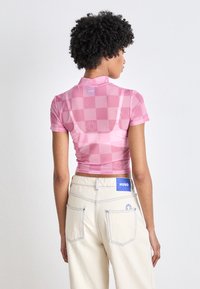 Rozā īsā krekla augšdaļa ar rūtainu rakstu un īsām piedurknēm, apvienota ar off-white augstās jostasvietas biksēm, kurām ir zilas šuves un logo.