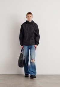 Zwarte windbreaker jas met rits, elastische manchetten en een capuchon. Paar vervaagde blauwe jeans met scheuren, zwarte schoenen en een donkere tote bag.