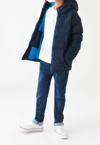 Marineblå pufferjakke med hætte, der har en lys blå indvendig, sammen med en hvid t-shirt, blå denimjeans og hvide sneakers.