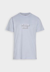 Camiseta de algodón azul claro de manga corta, con un logo en relieve en blanco y plata que dice "GUESS 1981" en el centro. Cuello redondo, sin estampados.