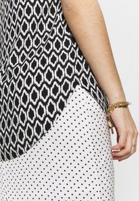 Haut noir et blanc à motif géométrique, avec une jupe à pois blancs contrastants à l'ourlet. Accent en chaîne dorée au poignet.