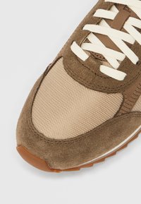 Smeđa tenisica s teksturiranim gornjim dijelom od brušene kože i bež mrežastim panelima, s bijelim vezicama i smeđim gumiranim potplatom.