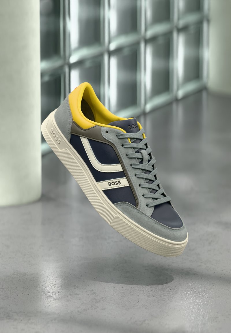 Σκούρο μπλε και γκρι αθλητικό παπούτσι με κίτρινες λεπτομέρειες, λευκές ρίγες και υλικά με υφή· διαθέτει λαστιχένια σόλα και επίπεδα κορδόνια.