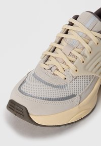 Beżowe i szare sportowe sneakersy z siateczkową cholewką, beżowym sznurówkami i gumową podeszwą, z podkreśleniem przodu i szczegółów sznurowania.