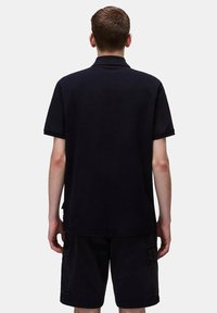 Polo nero in un tessuto a trama, maniche corte e colletto con tre bottoni, abbinato a pantaloni cargo neri, visto da dietro.