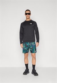 Zwarte lange mouwen shirt, groen en blauwe camouflage shorts, zwarte sneakers en zwarte sokken. Model met donkere zonnebril.