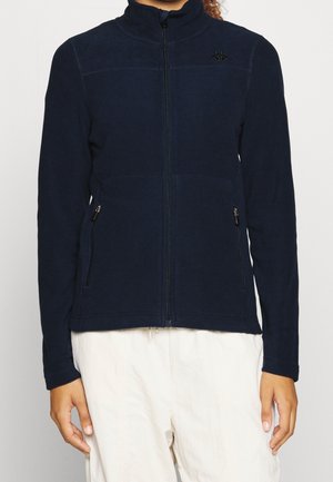 Personne portant une veste polaire bleu marine zippée avec des poches zippées et un pantalon blanc, montrée du cou aux hanches sur un fond uni.