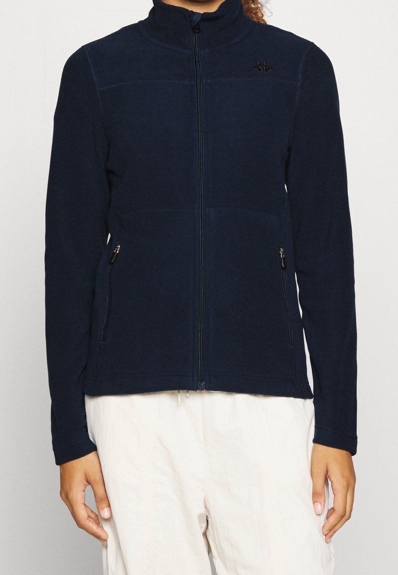 Personne portant une veste polaire bleu marine zippée avec des poches zippées et un pantalon blanc, montrée du cou aux hanches sur un fond uni.