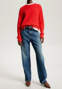 Personne portant un pull en maille rouge, un jean bleu avec une ceinture noire, et des chaussures marron à bout pointu, se tenant contre un fond neutre.