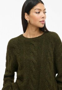 Austeres džemperis tumši zaļā krāsā ar kabeli rakstiem, apaļu dekoltē un brīvu piegriezumu. Mīksts, teksturēts audums. Garas piedurknes, ikdienišķs dizains.