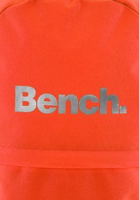 Narancssárga anyagú hátizsák, amelyen egy feltűnő ezüst "Bench." logó található, sima felülettel és letisztult vonalakkal. Az éleken varrott részletek.