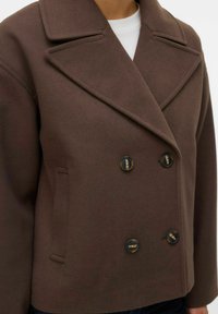 Brūns dubultpogājams žakete no teksturēta auduma, ar lielām melnām pogām un izgrieztām atlokiem, sānu kabatām un piegulošu siluetu.