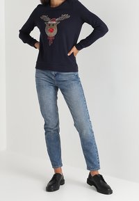 Osoba oblečená v tmavě modré mikině s motivem soba s červeným nosem, v světle modrých džínách a černých šněrovacích botách, stojící s rukama v bok.