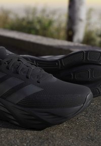 Close-up of black running shoes with textured soles placed outdoors on pavement near a blurred background of plants and a pole.
