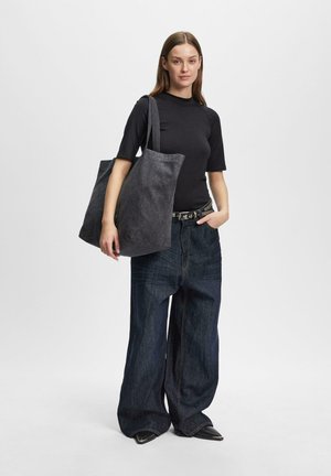 Junge Frau in schwarzem Oberteil und weitem, dunklem Jeansstoff, trägt große dunkle Einkaufstasche auf der Schulter, steht vor einfarbigem Hintergrund.