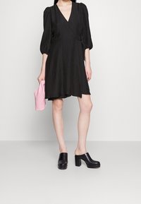 Robe enveloppante noire en tissu tissé avec un décolleté en V, des manches bouffantes, une fermeture à nouer et une forme en A ; associée à des sabots noirs à plateforme et à une pochette rose.