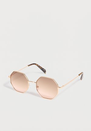 Osemuholníkové slnečné okuliare s tenkými zlatými kovovými rámami a hnedými gradientnými šošovkami na bielom pozadí.