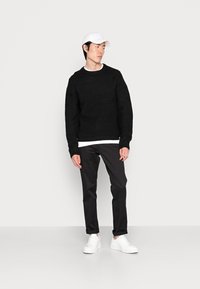 Maglione nero a maglia con motivo testurizzato, camicia bianca a girocollo sotto, pantaloni neri e sneakers bianche, completati da un cappellino bianco.