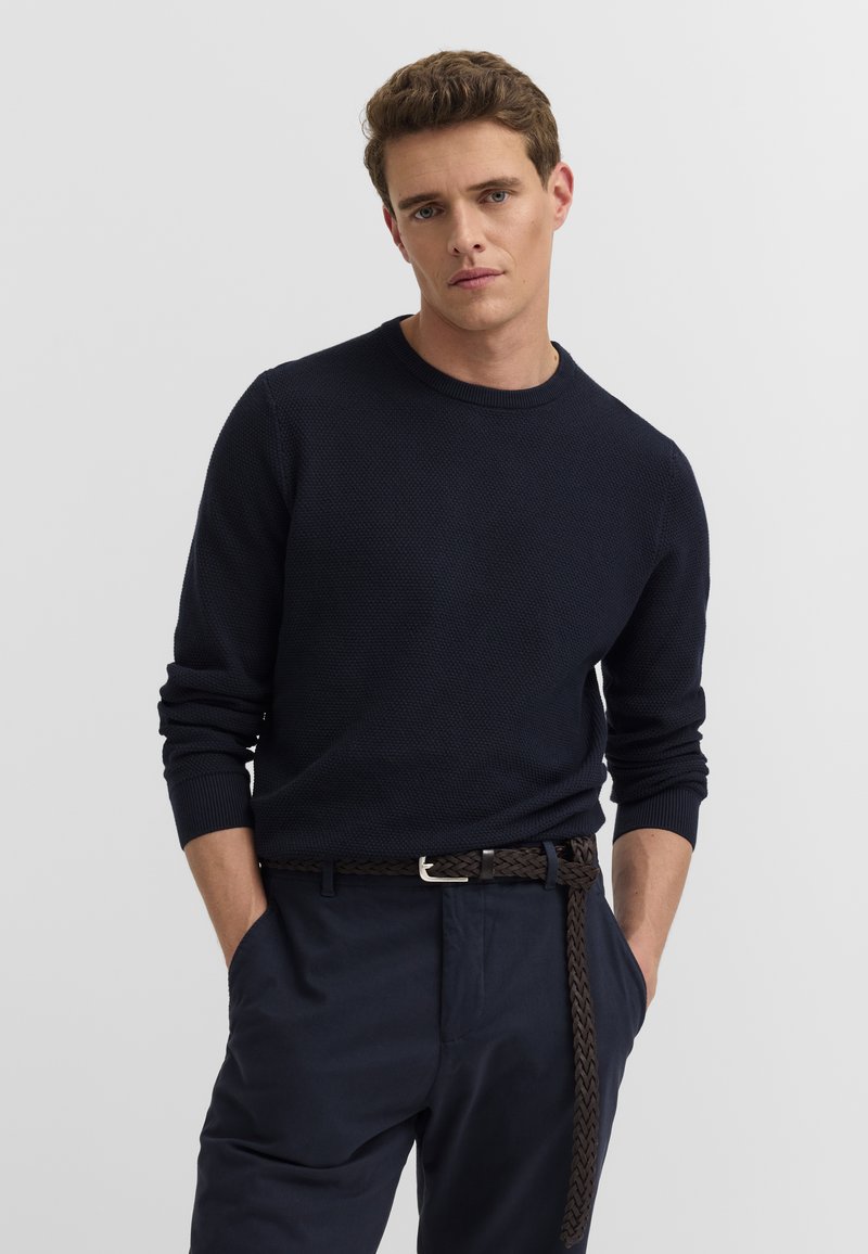 Junger Mann trägt einen dunkelblauen strukturierten Pullover und dunkelblaue Hose mit geflochtenem Gürtel, Hände in den Taschen, nach vorne blickend vor einem einfarbigen Hintergrund.