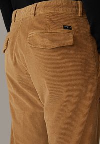 Close-up van bruin corduroy broek met achterzak en klein zwart merkje, gedragen met een zwart top.