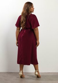 Burgundy, polvenpituisessa mekossa on lyhyet, liehuvat hihat; vyötärö on kireä ja pinta on sileä ja kiiltävä; yhdistettynä kultaisiin nilkkavöihin.