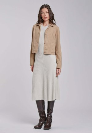 Vrouw draagt een beige cropped jack over een lichtgrijze midijurk met donkerbruine kniehoge laarzen met hoge hakken, staand tegen een effen achtergrond.
